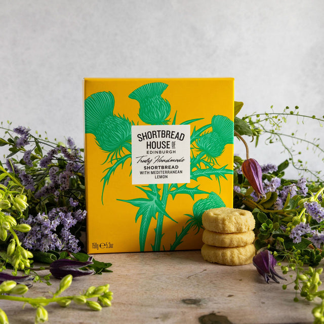 A yellow box of Shortbread House of Edinburgh Lemon Handmade Shortbread cookies stands on a rustic wooden surface surrounded by purple flowers, with three stacked cookies in front of the box.