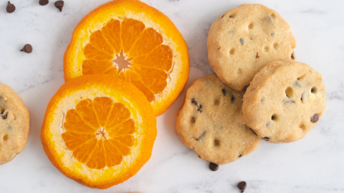 Chocolate chip shortbread arranged on a white marble surface next to two sliced orange halves with scattered chocolate chips.