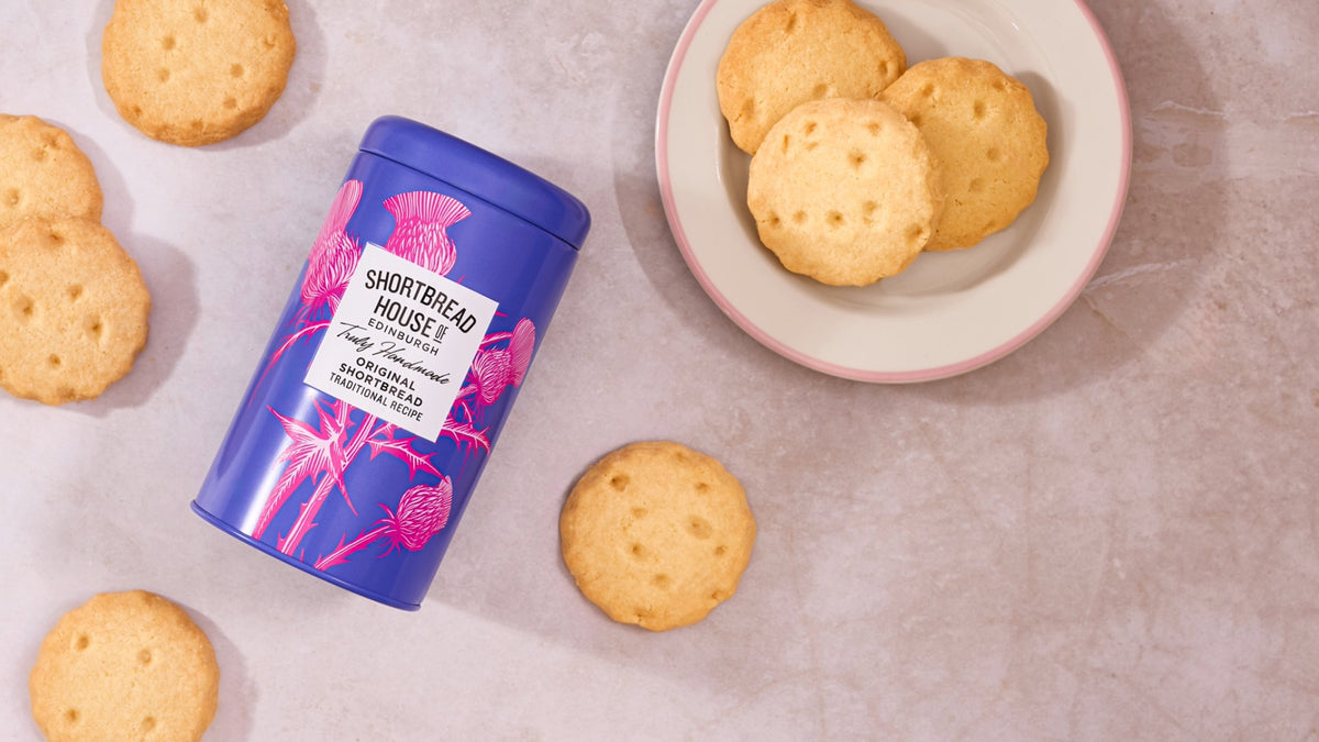 Shortbread House biscuits are shown with their purple and pink branded tin on a light textured surface.