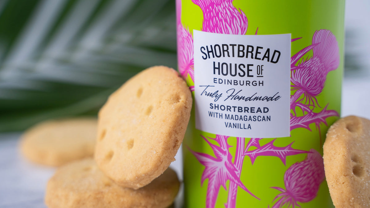A close-up of a bright green tin of Shortbread House of Edinburgh vanilla shortbread cookies, surrounded by several golden-brown round shortbread cookies on a light surface with a blurred green leaf in the background.