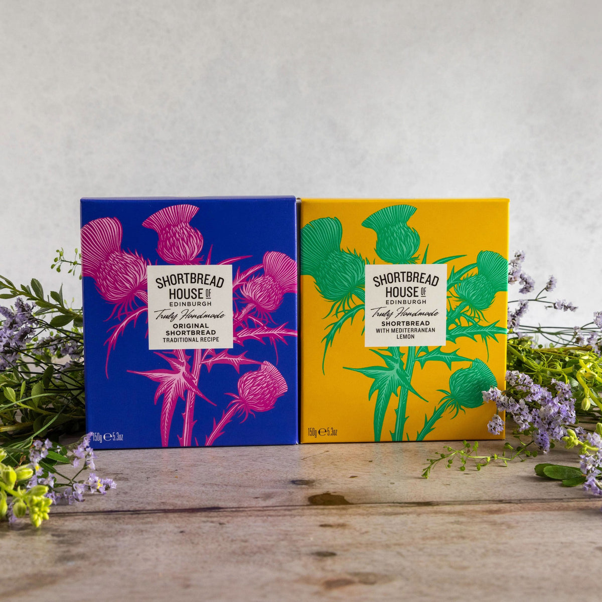 Two boxes of Shortbread House Shortbread, one blue with pink thistle illustrations and one yellow with green thistle illustrations, displayed on a rustic wooden table with purple flowers.