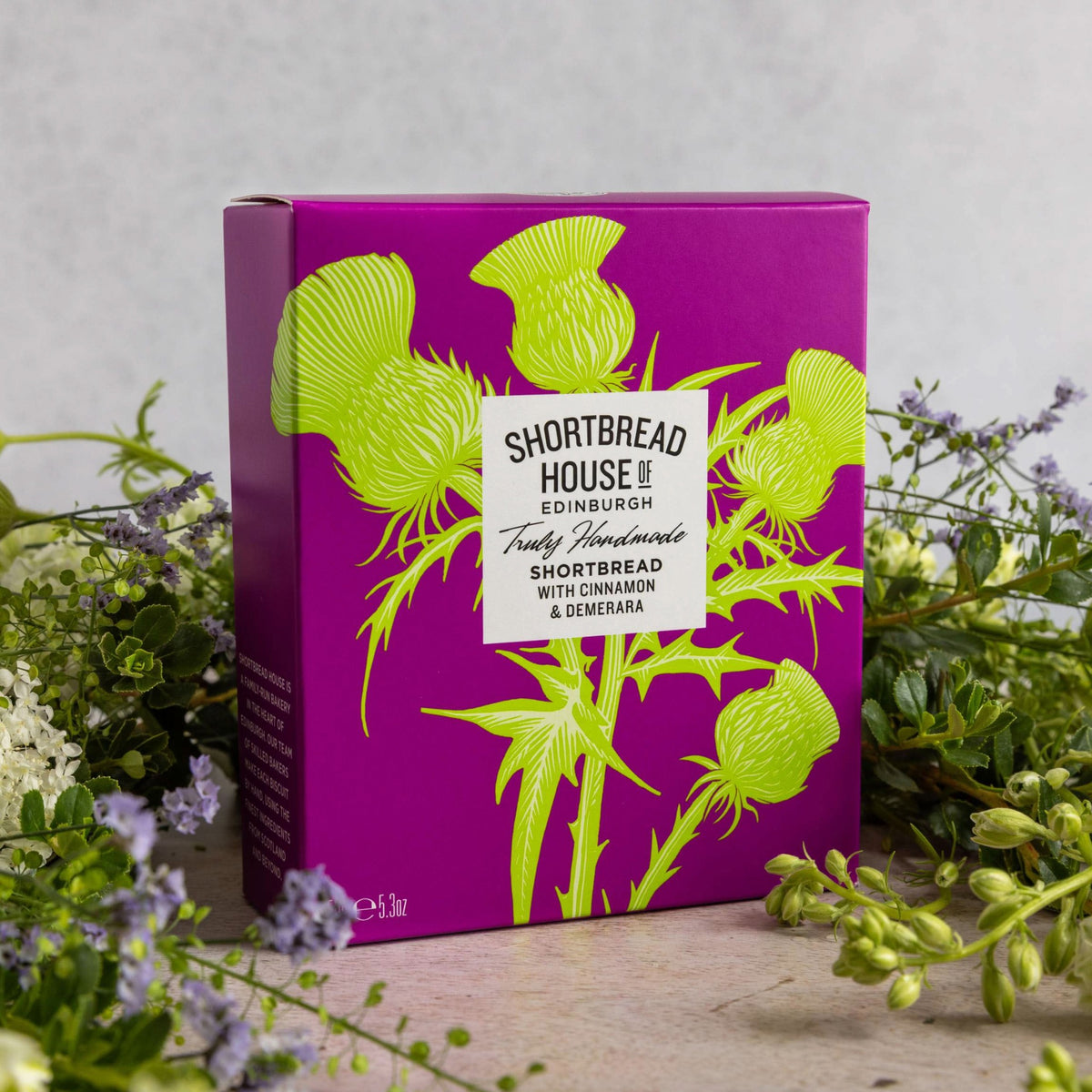 A purple and green Shortbread House of Edinburgh box surrounded by a floral arrangement on a natural wooden surface