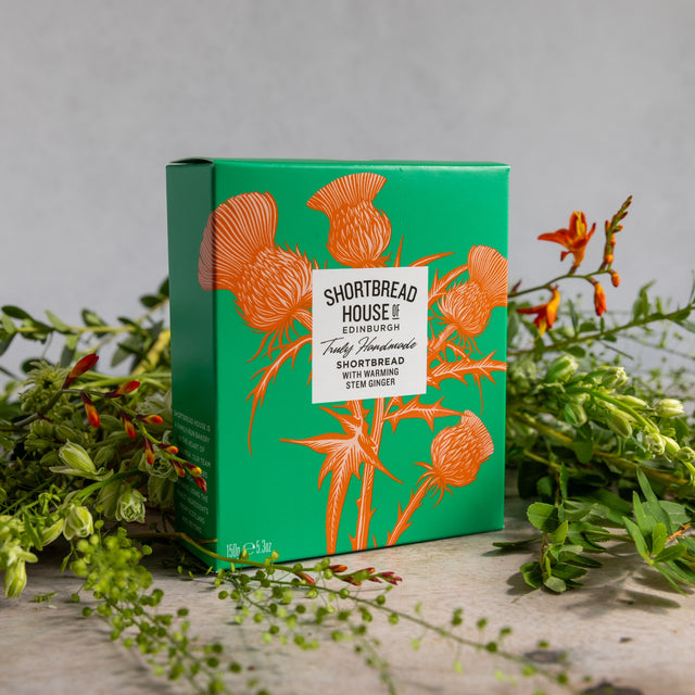 A green rectangular product box with orange thistle illustrations and a white label reading 'SHORTBREAD HOUSE OF EDINBURGH' styled on a light surface with greenery around it.