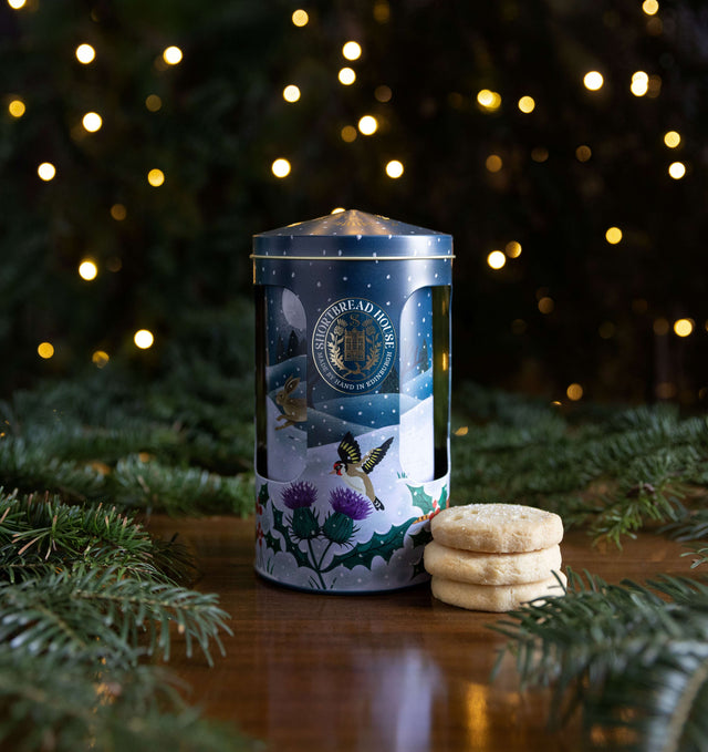 A navy Shortbread House musical tin with a Christmas scene on a surface with fir and three biscuits stacked in front.