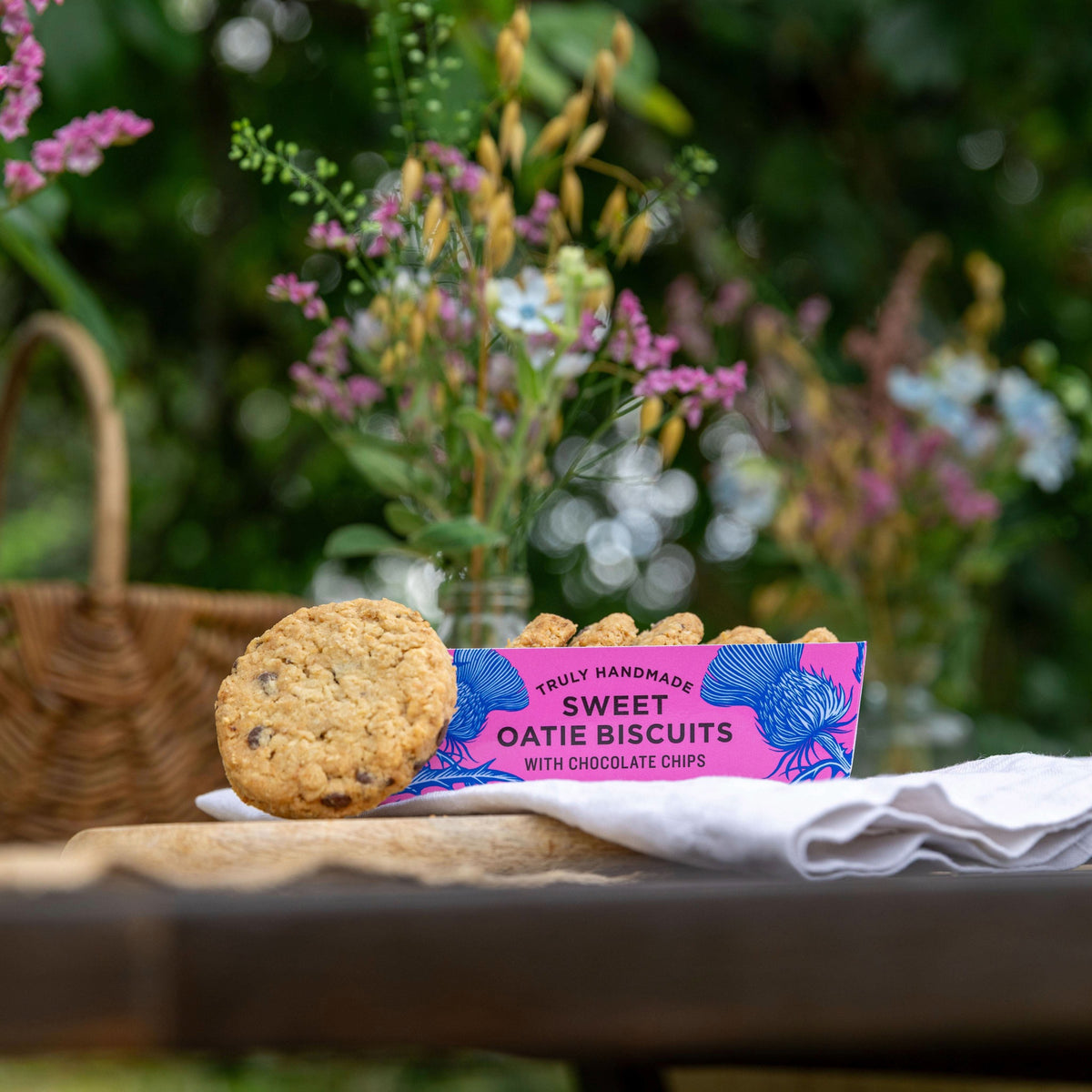A pink tray of sweet oatie biscuits, pictured on an outdoor tabletop, with a chocolate chip oatie leaning against the tray.