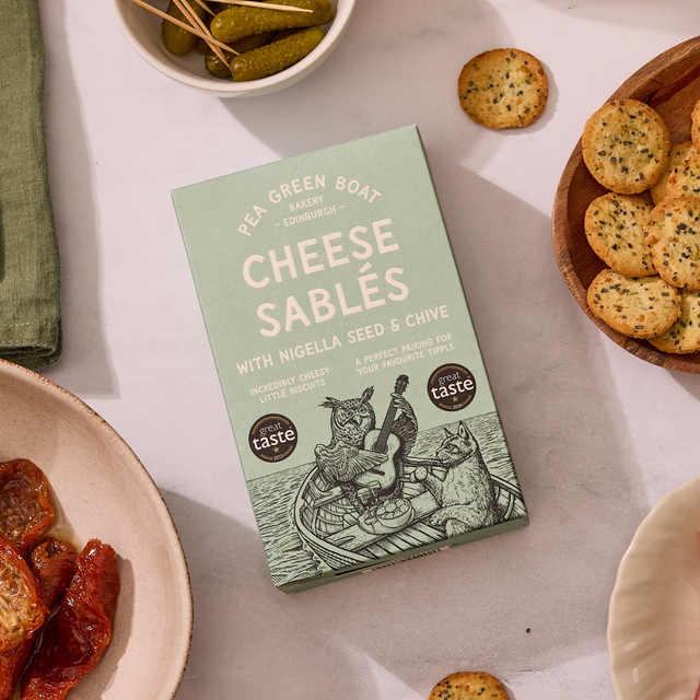 Box of Cheese Sablés with Nigella Seed & Chive on a table with sundried tomatoes and gherkins.