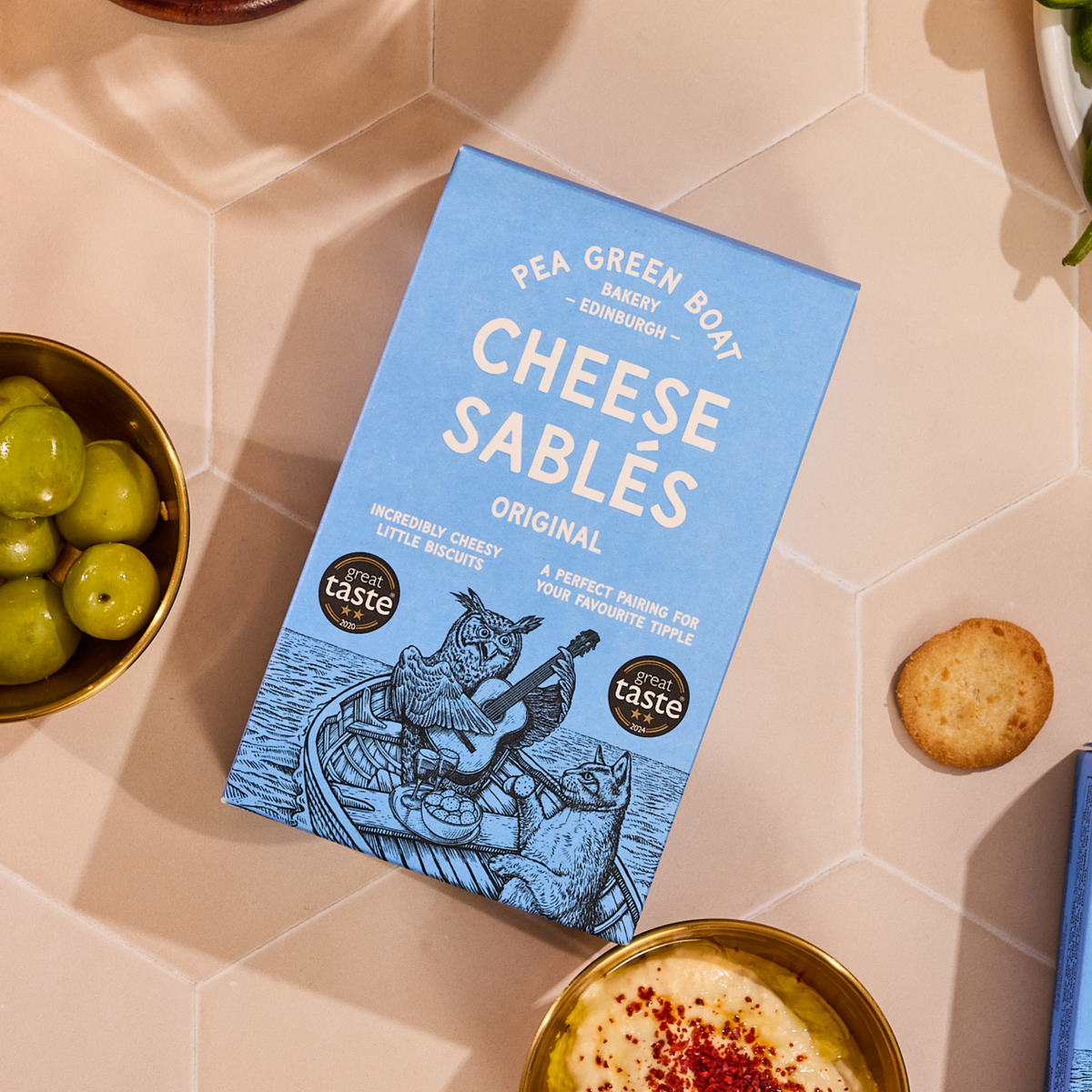 Packaging of Pea Green Boat Cheese Sablés on a tiled surface with snacks.