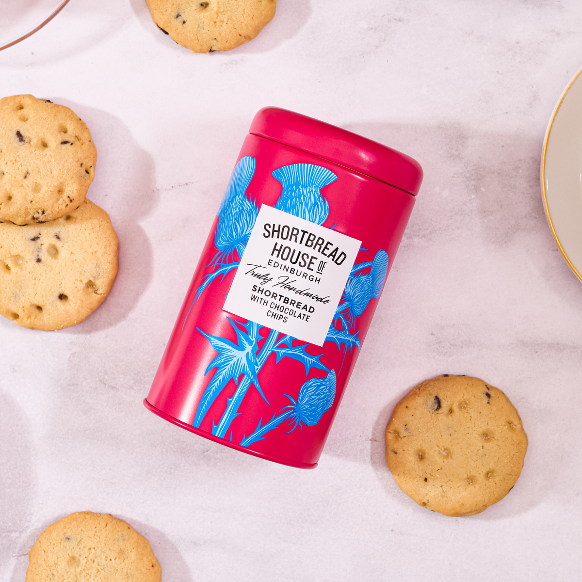"A pinkish red tin with blue thistle tin of Shortbread House of Edinburgh Original Traditional Recipe shortbread, surrounded by round golden biscuits with chocolate chips on a light stone surface with a plate of biscuits in the background."