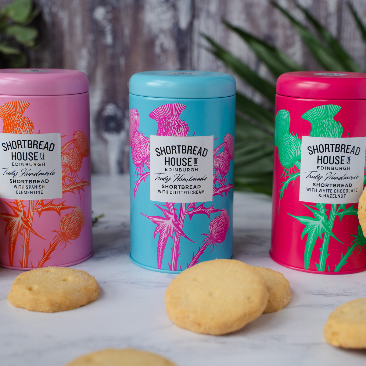 Three colorful tins of Shortbread House of Edinburgh shortbread biscuits on a marble surface with biscuits in front.