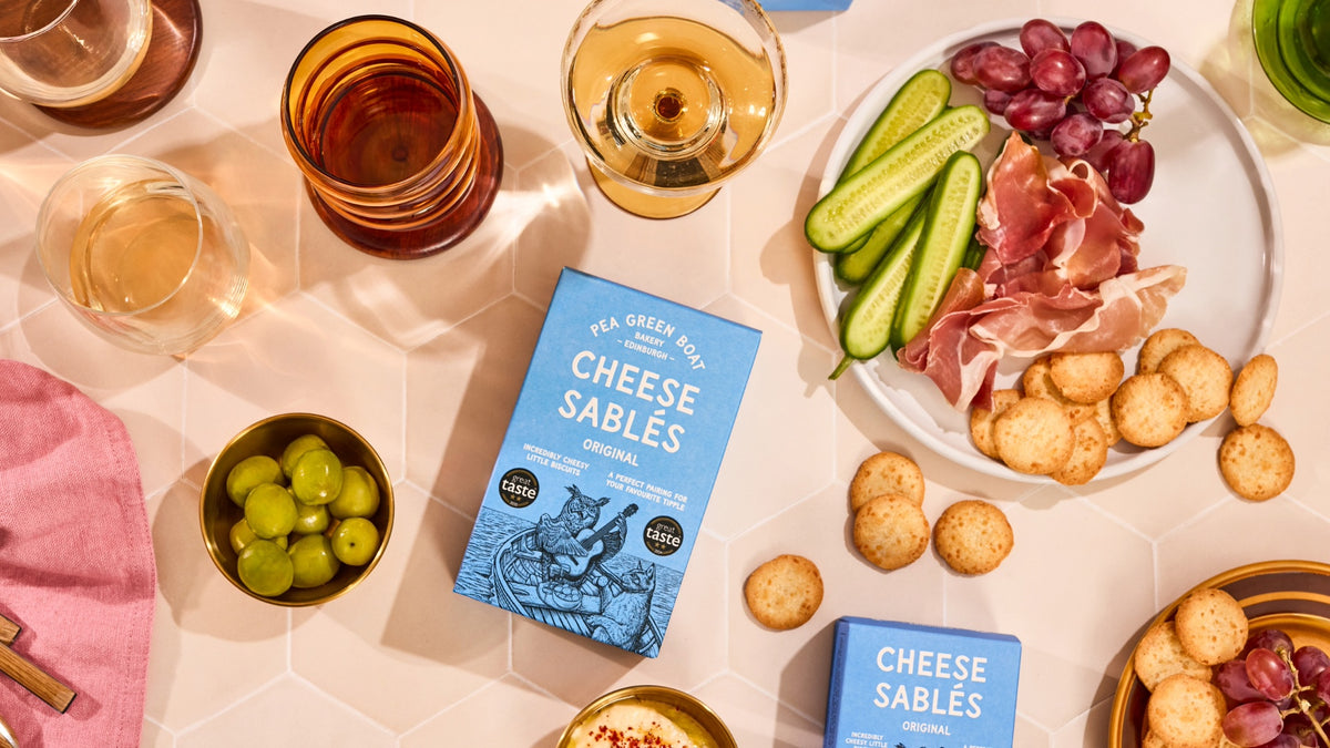 A styled tabletop flat lay featuring a box of Cheese Sablés. The blue packaging is surrounded by a charcuterie board with crackers, prosciutto, cucumbers, grapes, and glasses of wine on a light pink hexagonal surface.