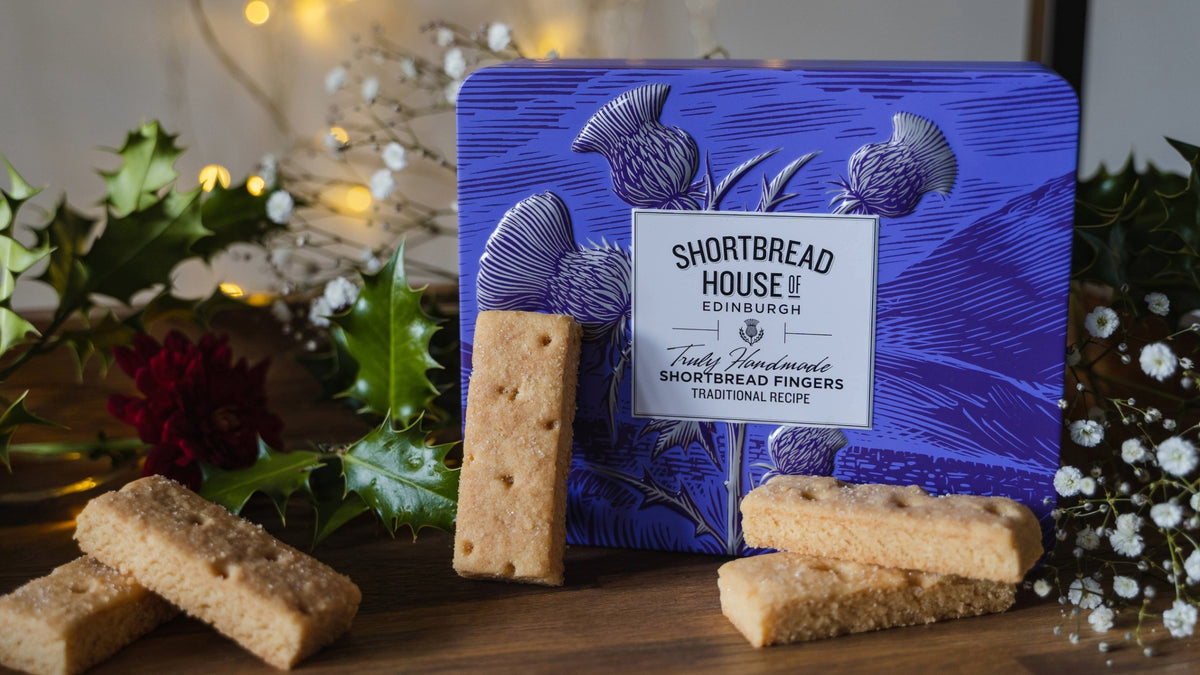 Shortbread House Traditional Shortbread Fingers displayed with their blue illustrated tin packaging on a wooden surface, surrounded by holly leaves, white flowers, and warm string lights.