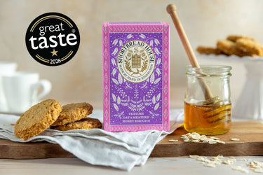 A box of Shortbread Biscuits stands on a wooden board with cookies, oats, a jar of honey, and a honey dipper in a styled kitchen scene. A black circular badge in the upper left reads 'great taste 2026'.