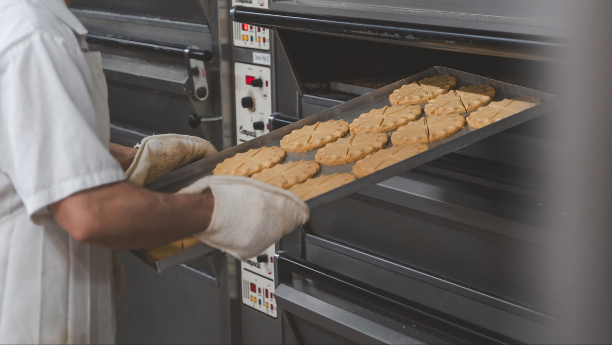 A baker wearing a white uniform and oven mitts slides a tray of freshly baked cookies into a commercial oven.