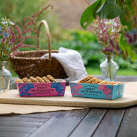 Two boxes of Sweet Oatie Biscuits with Chocolate Chips are displayed on a wooden board on an outdoor table. The background features a wicker basket, white cloth, and glass vases with green foliage and pink flowers.