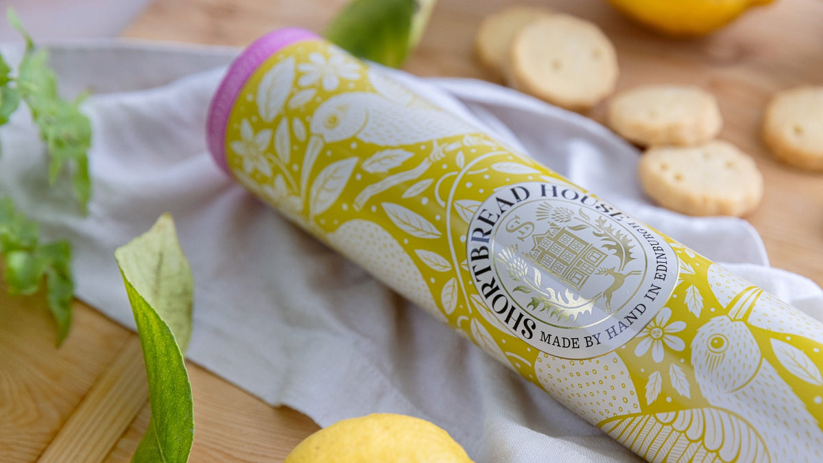 A yellow tin of lemon shortbread at an angle on a wooden board surrounded by lemons and biscuits.