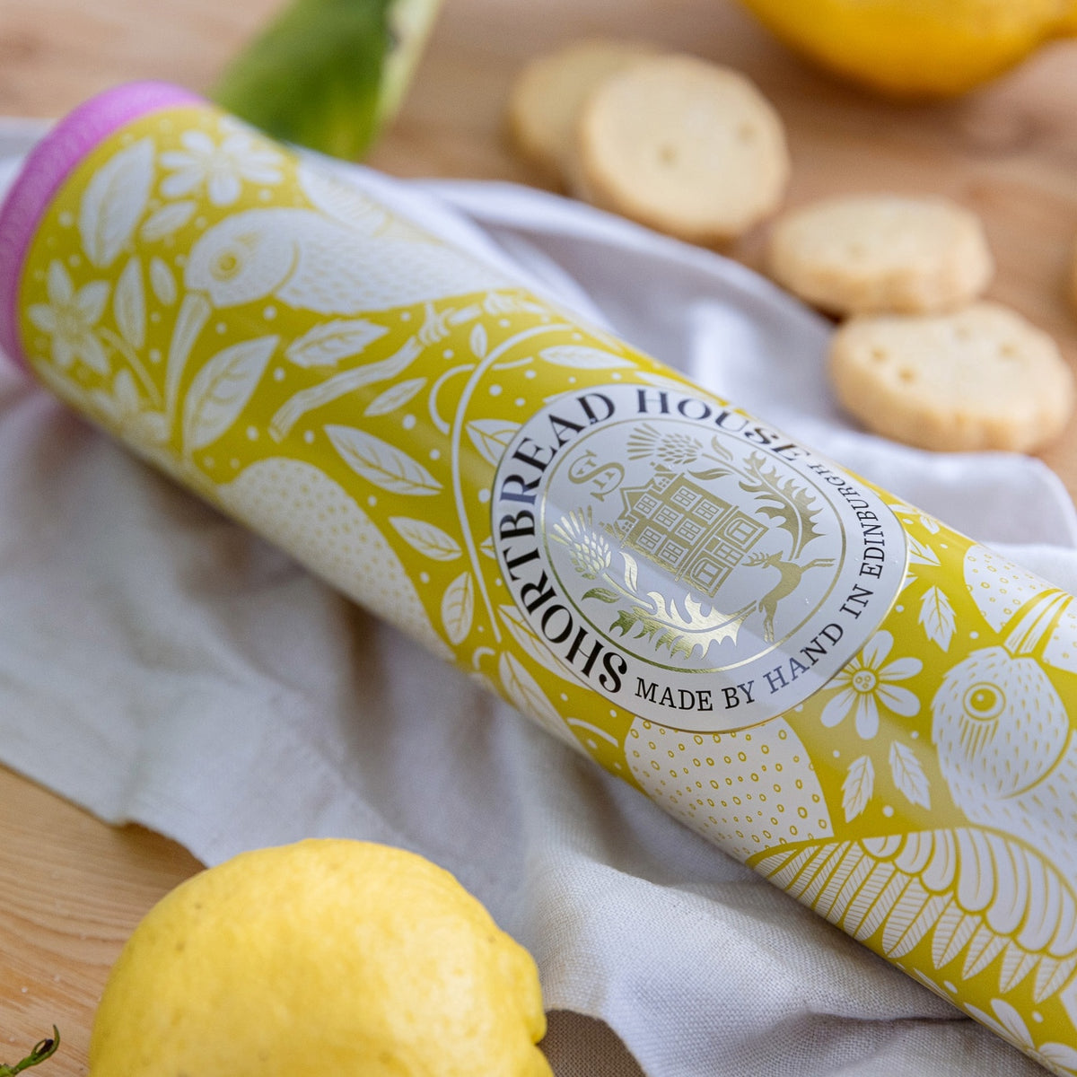 Tin of lemon shortbread on a wooden board with lemons and biscuits.