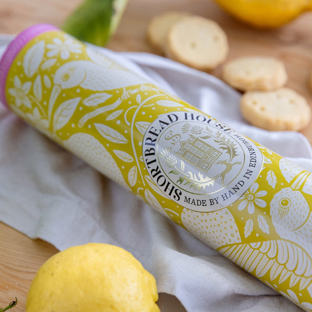 Tin of lemon shortbread on a wooden board with lemons and biscuits.