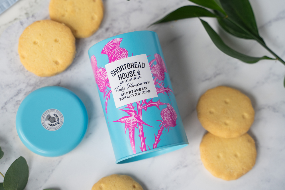 A blue tin of shortbread with pink thistles on a marble surface, surrounded b shortbread biscuits and green leaves.