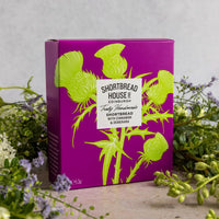 A purple and green Shortbread House of Edinburgh box surrounded by a floral arrangement on a natural wooden surface