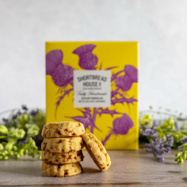 A stack of golden-brown shortbread cookies with chocolate chips stands in front of a bright yellow box decorated with purple thistle illustrations. The scene is styled on a wooden surface with soft green and purple flowers in the background.