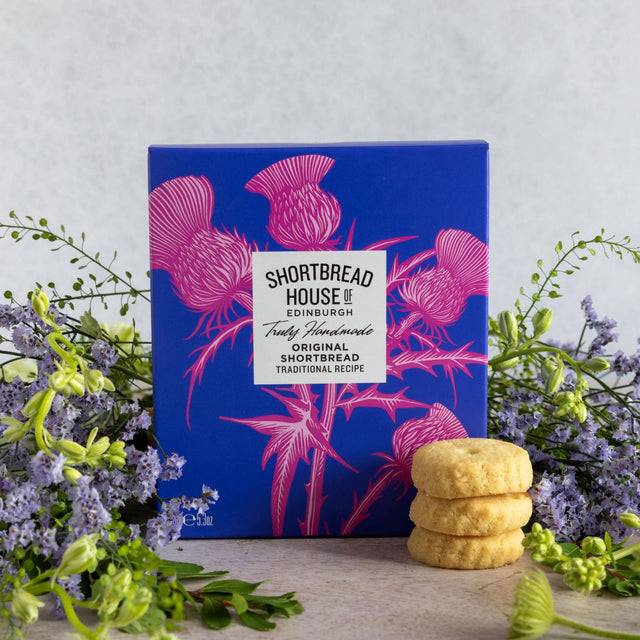 A blue box of Shortbread House of Edinburgh Original Shortbread cookies, decorated with pink thistle illustrations and a white brand label. The box is styled on a light surface with green and purple flowers on both sides and three golden-brown shortbread cookies stacked in front.