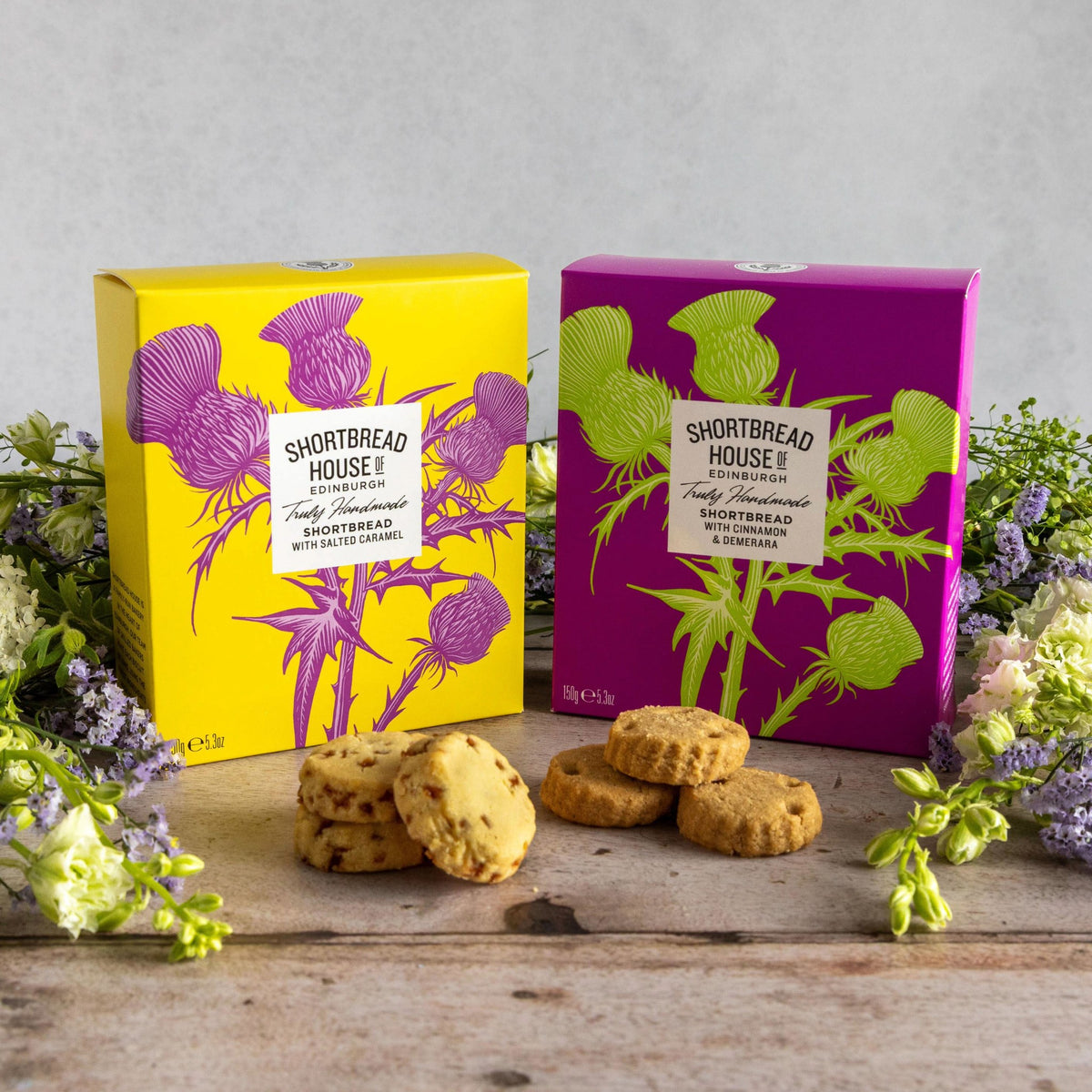 Two Shortbread House of Edinburgh boxes with colourful thistle designs. The boxes are pictures surrounded by a floral arrangement with salted caramel biscuits and cinnamon and demerara biscuits sitting in front.
