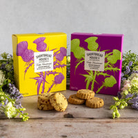 Two Shortbread House of Edinburgh boxes with colourful thistle designs. The boxes are pictures surrounded by a floral arrangement with salted caramel biscuits and cinnamon and demerara biscuits sitting in front.
