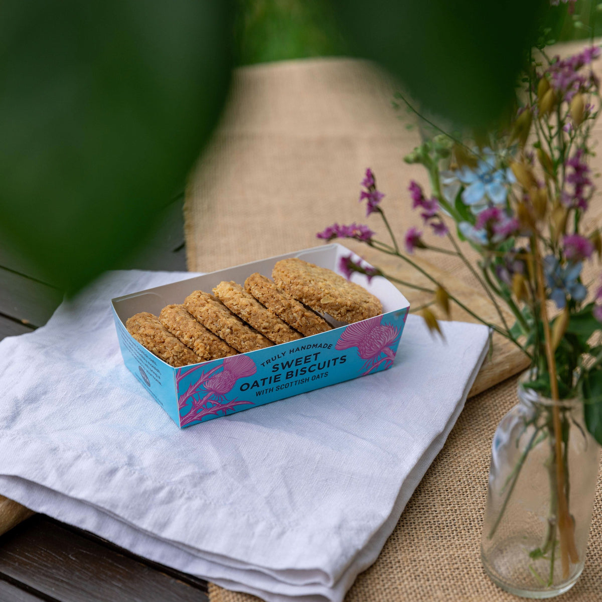 A box of Sweet Oatie Biscuits sits on a white cloth on a burlap surface, with a glass jar of flowers nearby and soft green leaves framing the top of the scene.