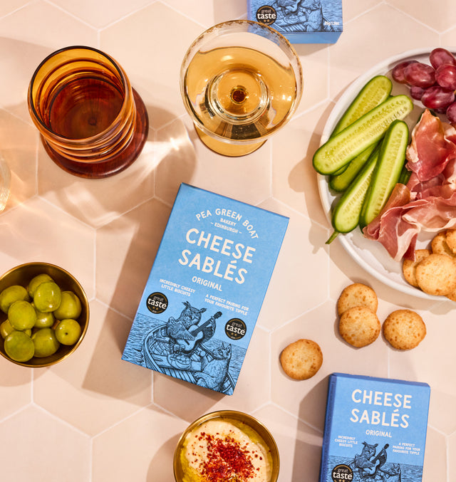 Overhead view of a charcuterie-style spread on a tiled surface, featuring boxes of cheese sablé biscuits, a glass of white wine, olives, sliced cucumber, grapes and cured meats arranged neatly on a plate.