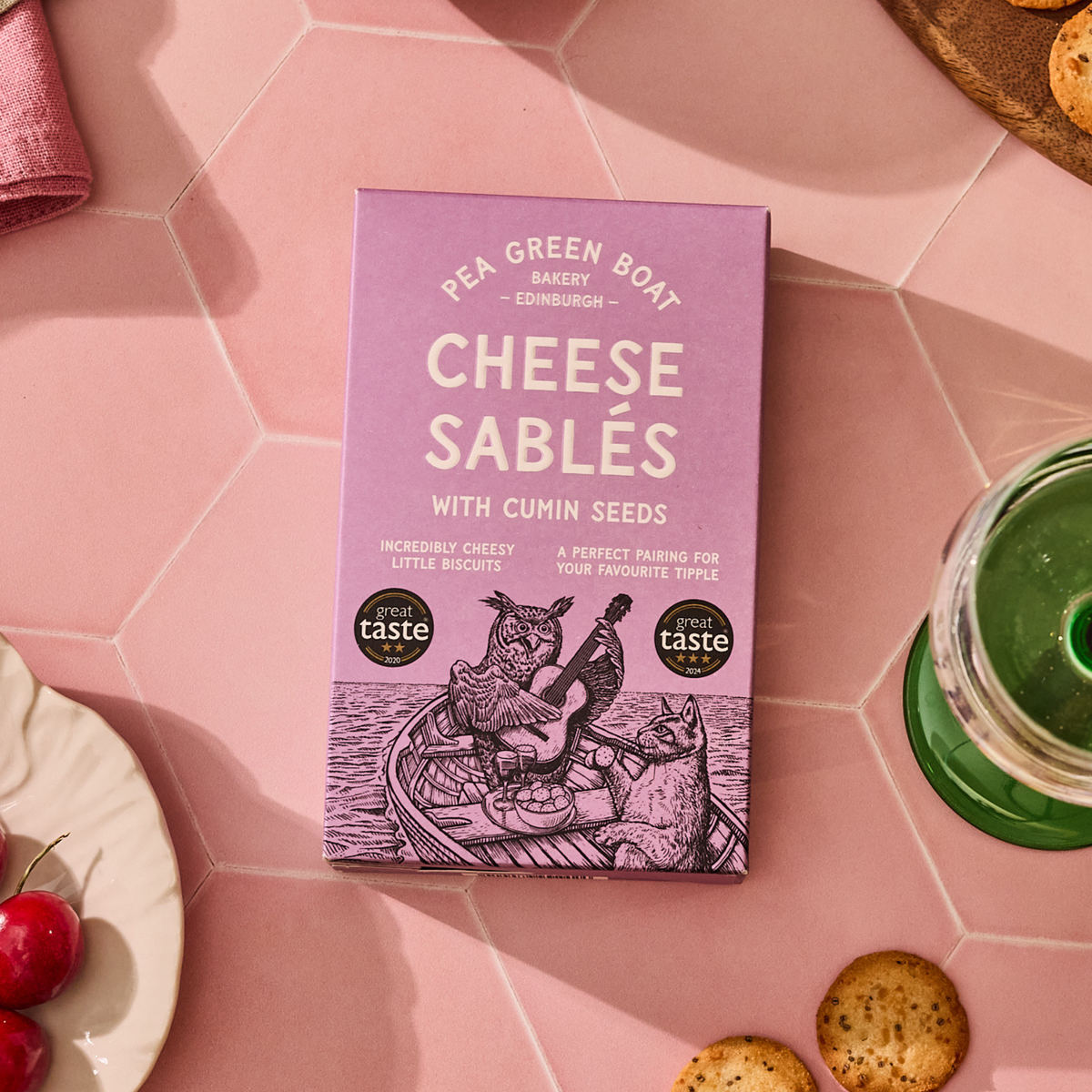 Purple box of 'Cheese Sablés with Cumin Seeds' from Pea Green Boat Bakery on a pink tiled surface.
