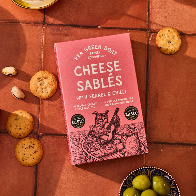 Red box of Cheese Sables with fennel and chilli on a terracotta surface with biscuits and olives.