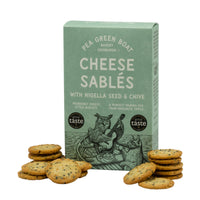 Green box of Pea Green Boat Cheese Sablés, surrounded by stacks of golden cheesy biscuits. The packaging features an illustrated owl playing a guitar in a boat.