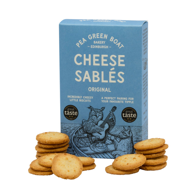Blue box of Pea Green Boat Cheese Sablés, surrounded by stacks of golden cheesy biscuits. The packaging features an illustrated owl playing a guitar in a boat.