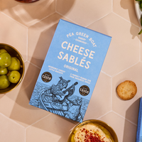 Packaging of Pea Green Boat Cheese Sablés on a tiled surface with snacks.