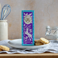 Purple shortbread box with a deer design on a table with biscuits and kitchen utensils