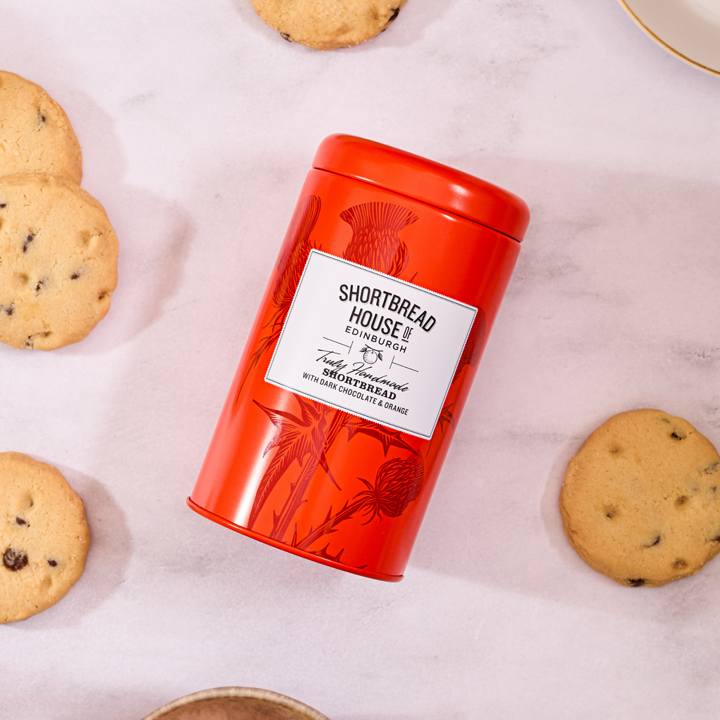 Red-ish orange tin labeled 'Shortbread House of Edinburgh' with biscuits around on a light surface