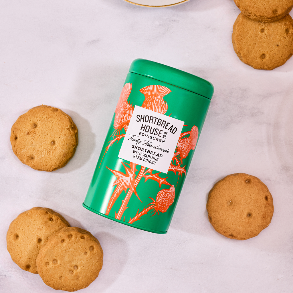 Green tin with orange thistle design of Shortbread House shortbread on a white surface with biscuits around it.