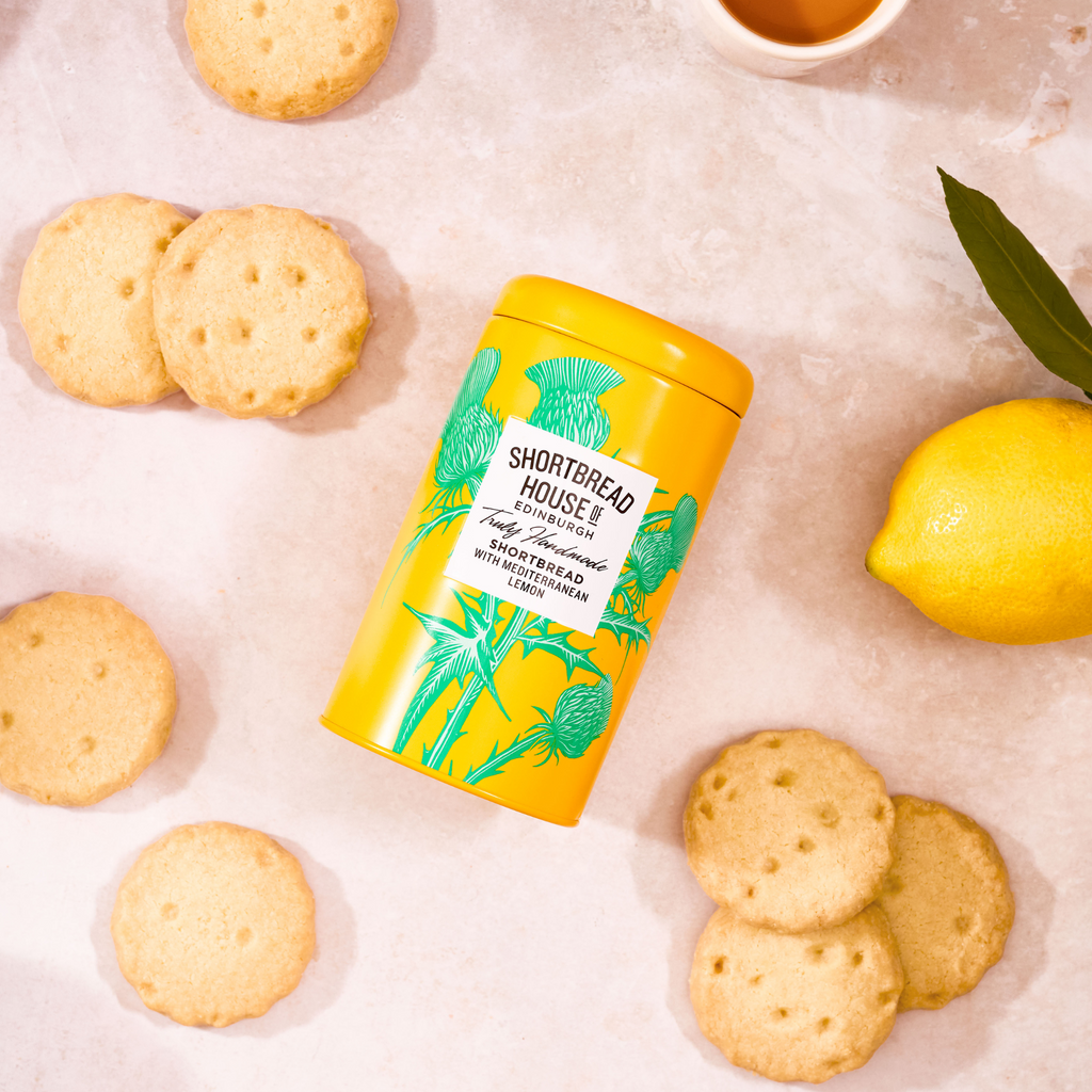 Yellow tin of Shortbread House lemon shortbread with round biscuits and a lemon on a light background