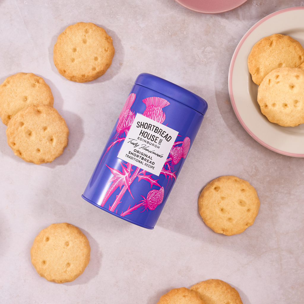 A purple tin of Shortbread House of Edinburgh Original Traditional Recipe shortbread, surrounded by round golden biscuits on a light stone surface with a plate of biscuits in the background.