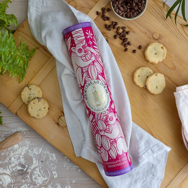 Pink and purple shortbread tin with decorative design on a wooden board with cookies and plants.