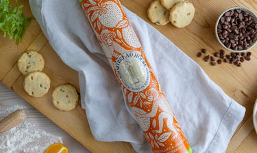Orange tin of shortbread with monkey and orange illustration on wooden chopping board with biscuits and chocolate chips.