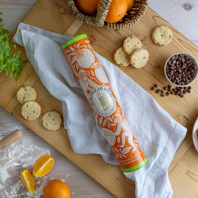 An orange shortbread tin with a monkey design on a wooden chopping board surrounded by cookies, oranges and chocolate