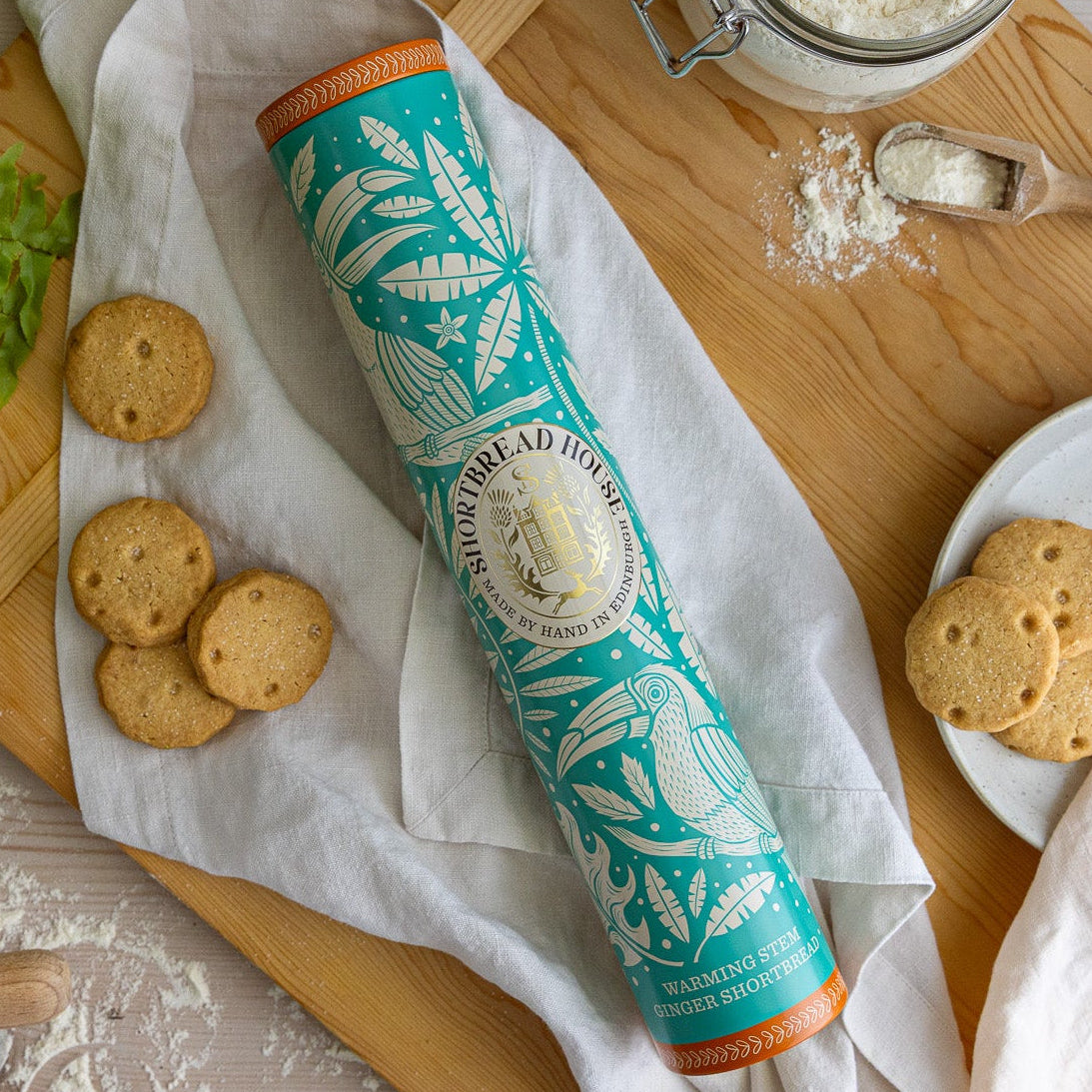 Tall tin with a blue toucan design on a chopping board with biscuits and flour