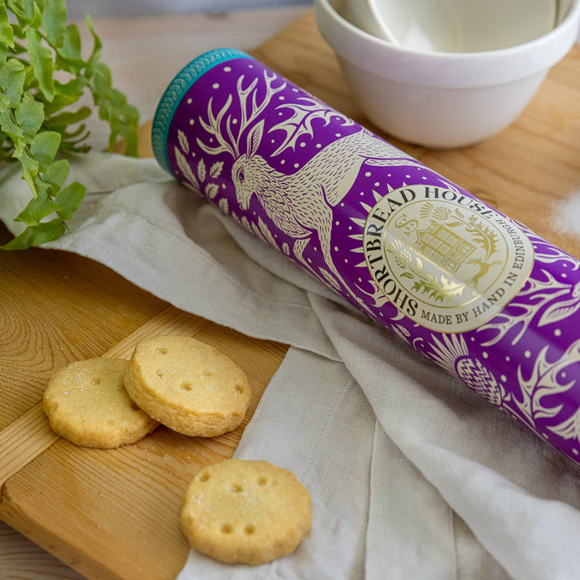 A purple shortbread tin with a stag design on a wooden chopping board with sugar, biscuits and greenery in the background.