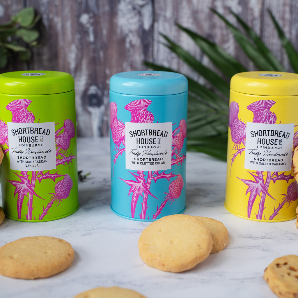 Three colorful tins of Shortbread House shortbread biscuits on a marble surface with biscuits around them.