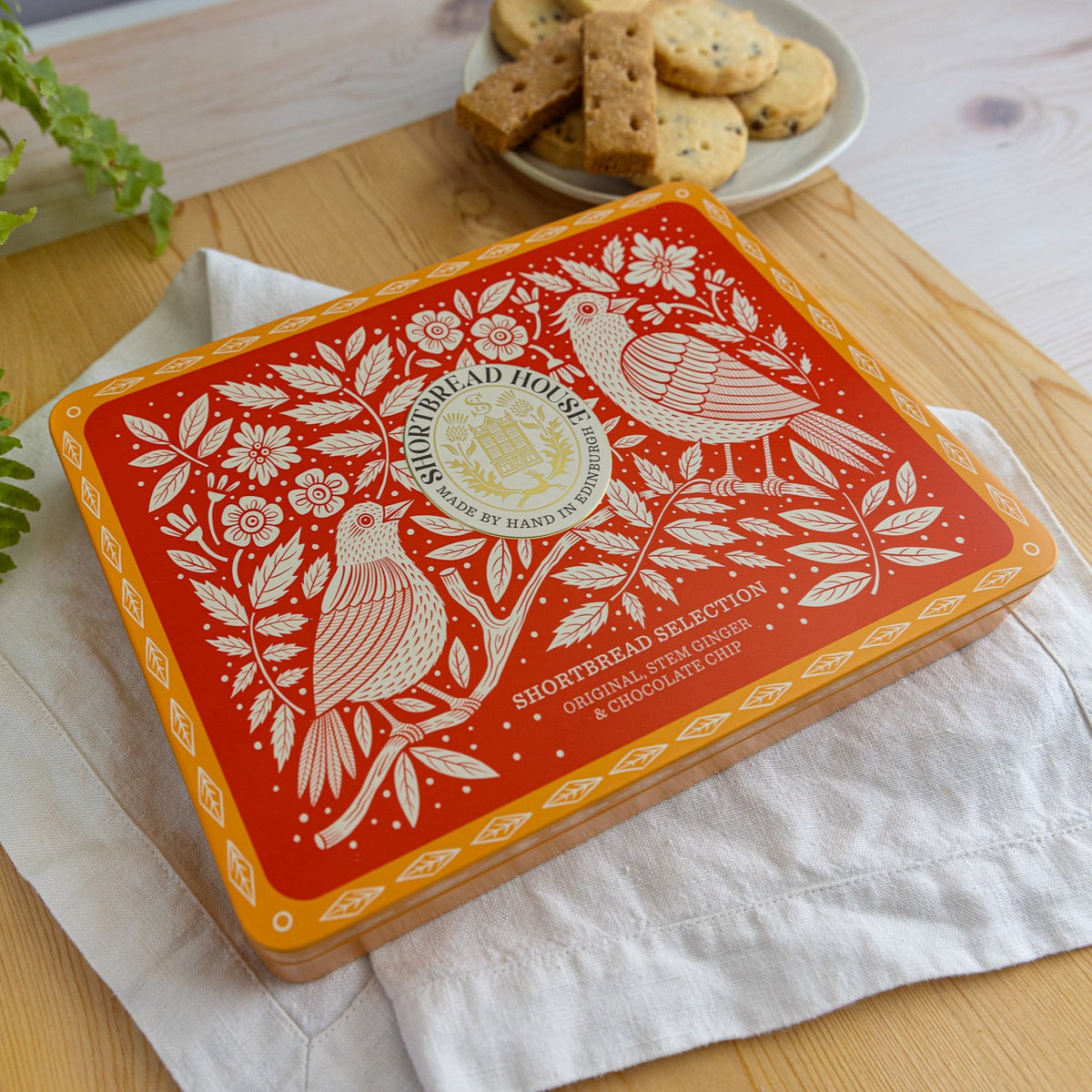 A rectangular tin of Shortbread House biscuits  sits on a white cloth over a wooden surface, with a plate of biscuits in the background.