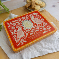 A rectangular tin of Shortbread House biscuits  sits on a white cloth over a wooden surface, with a plate of biscuits in the background.