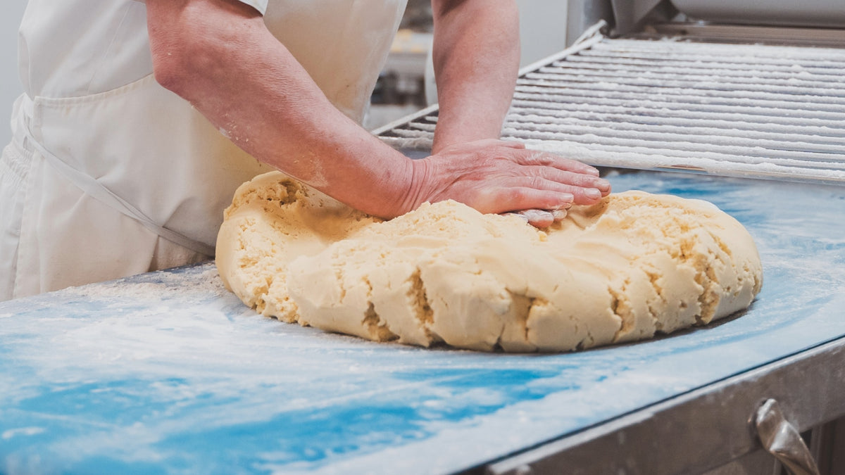 A close-up of a baker's hands pressing down a large mound of pale dough on a blue surface