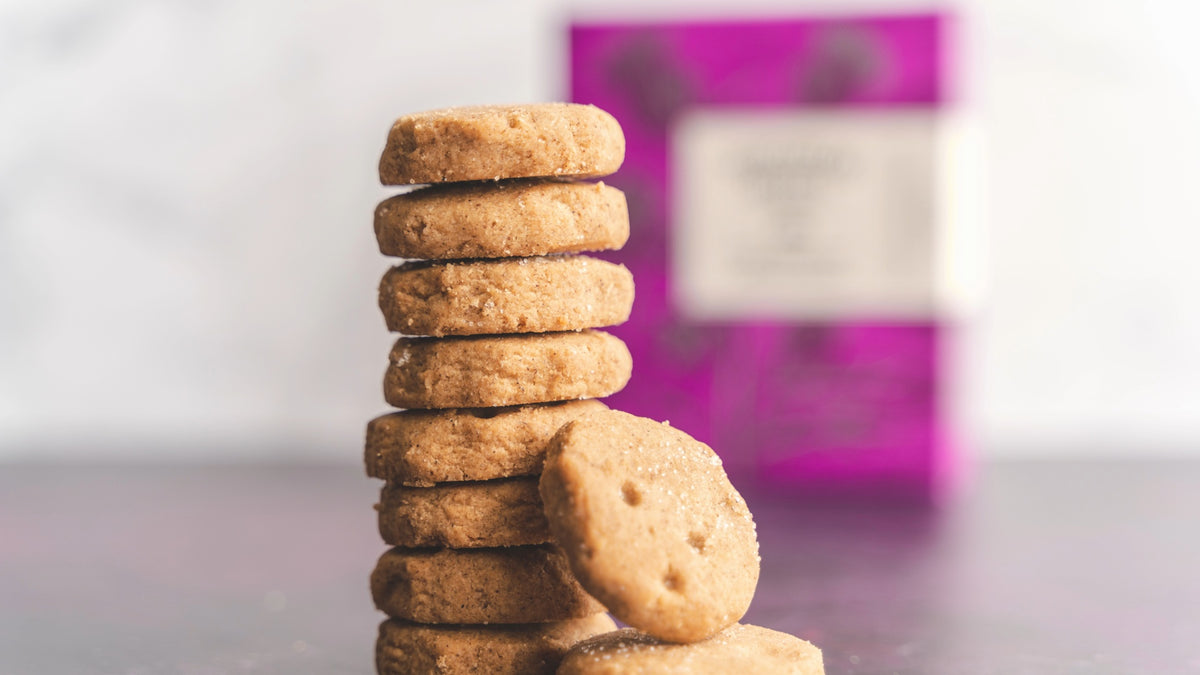 A stack of golden-brown shortbread cookies on a dark surface, with a blurred pink and white box in the background.