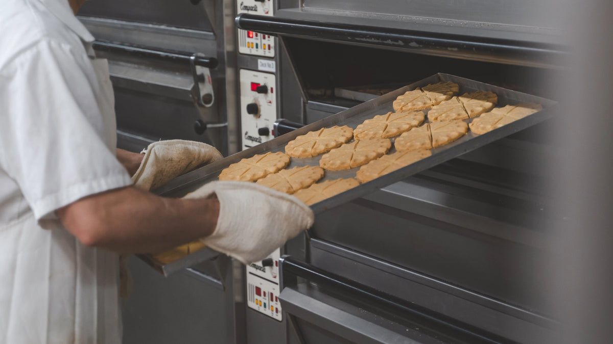 A baker wearing a white uniform and protective gloves slides a tray of freshly baked cookies out of a large commercial oven.